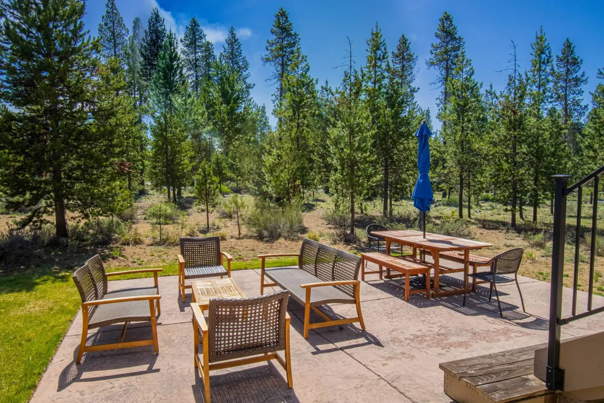 An inviting outdoor dining area set amidst tall trees, featuring a table with chairs and a vibrant blue umbrella. The space suggests a peaceful, natural environment ideal for picnics, outdoor meals, or casual gatherings. Sunlight filters through the trees, creating a tranquil yet welcoming mood.