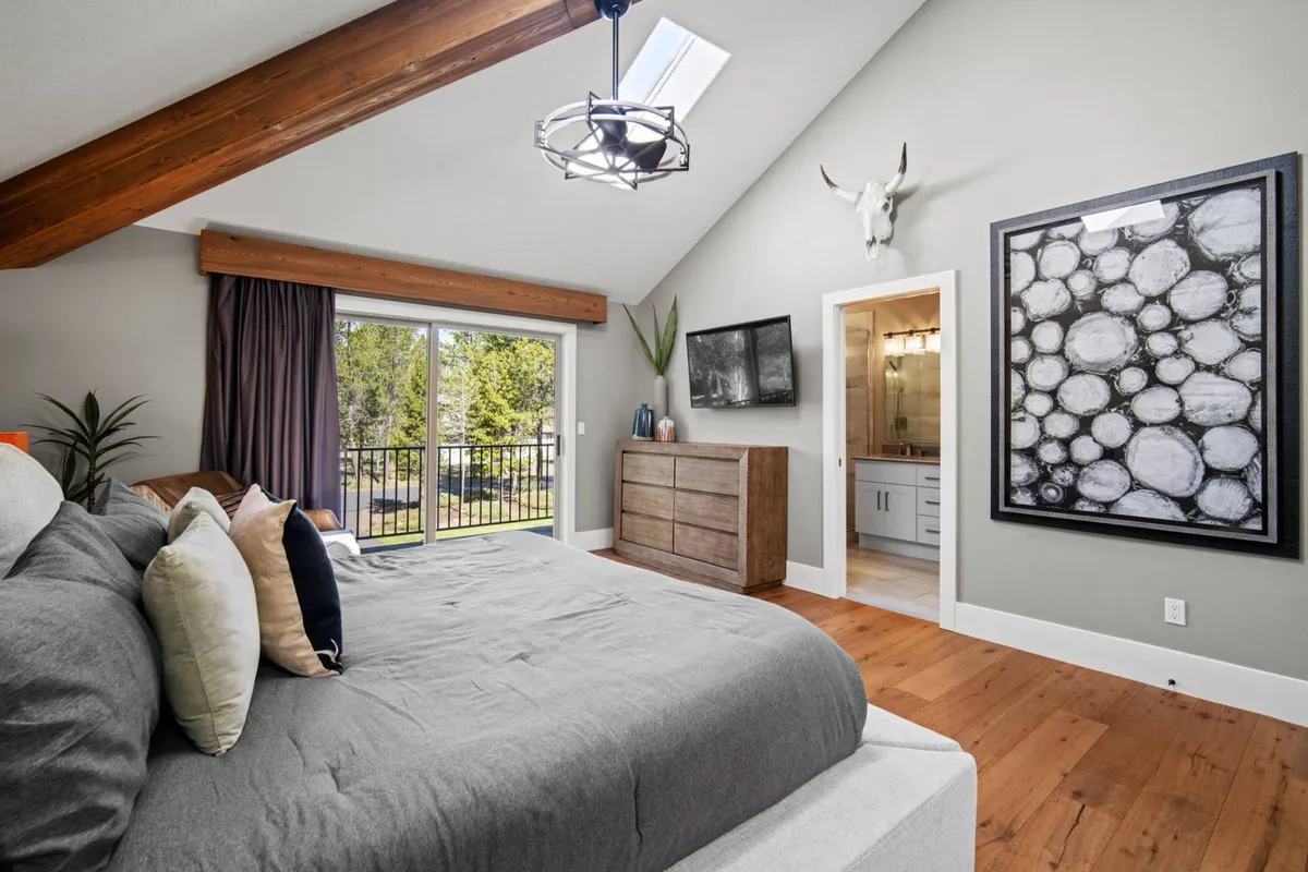 A spacious bedroom featuring a large bed adorned with decorative pillows, exposed wooden beams on the ceiling, a large window offering a scenic view, and tasteful wall art. The space feels light-filled and tranquil, ideal for relaxation.
