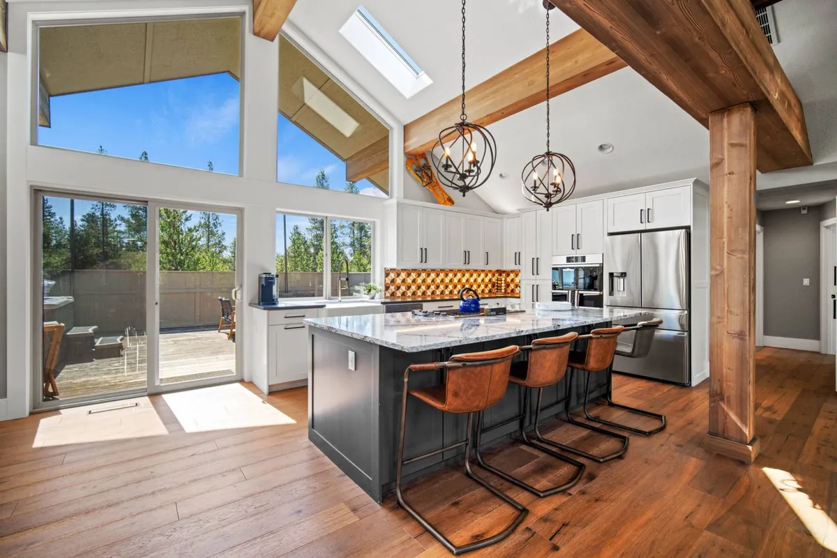A spacious, modern kitchen featuring a central island, bar stools, and large windows that flood the room with sunlight. Wooden beams add warmth and character, while the layout encourages socializing, cooking, and entertaining. The open-plan design emphasizes luxury, space, and comfort.
