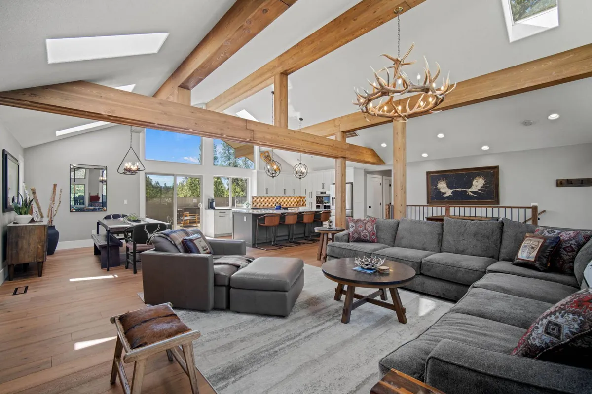 A wide-angle view of a spacious open-concept area, seamlessly integrating the living room, dining table, kitchen bar, and decorative antler chandelier. The scene displays ample plush seating, wooden beams, and a welcoming atmosphere designed for group gatherings and entertaining.
