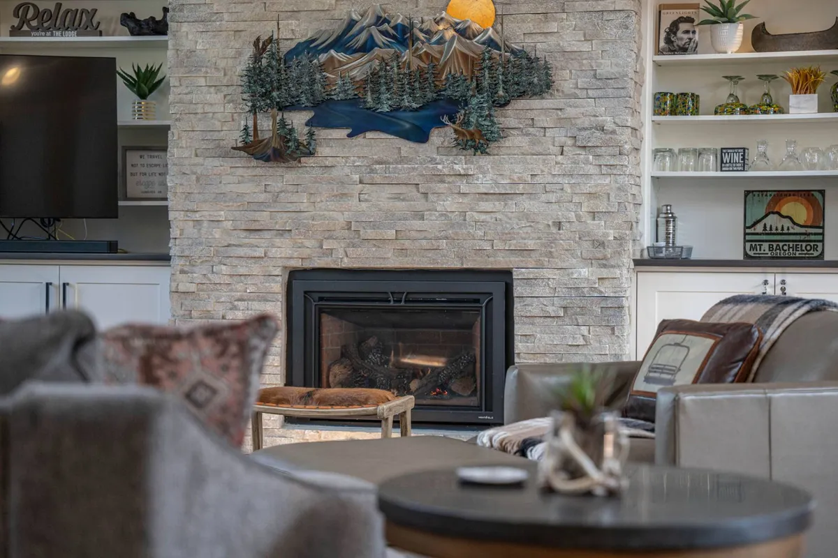 This cozy living room centers around a striking stone fireplace with a wooden mantel, flanked by comfortable seating and a mountain mural. The ambiance is warm and welcoming, suggesting evenings spent relaxing by the fire or sharing stories with friends and family.