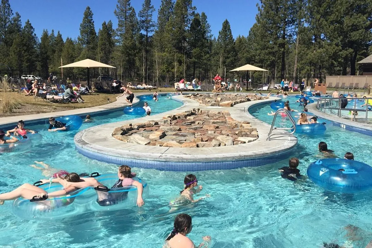 A sun-drenched exterior shot of a lazy river with people floating on tubes, sun umbrellas set up along the water's edge, and a rock formation that adds natural beauty. The surrounding area is lively, with families and groups enjoying leisurely water activities and socializing under the sun.