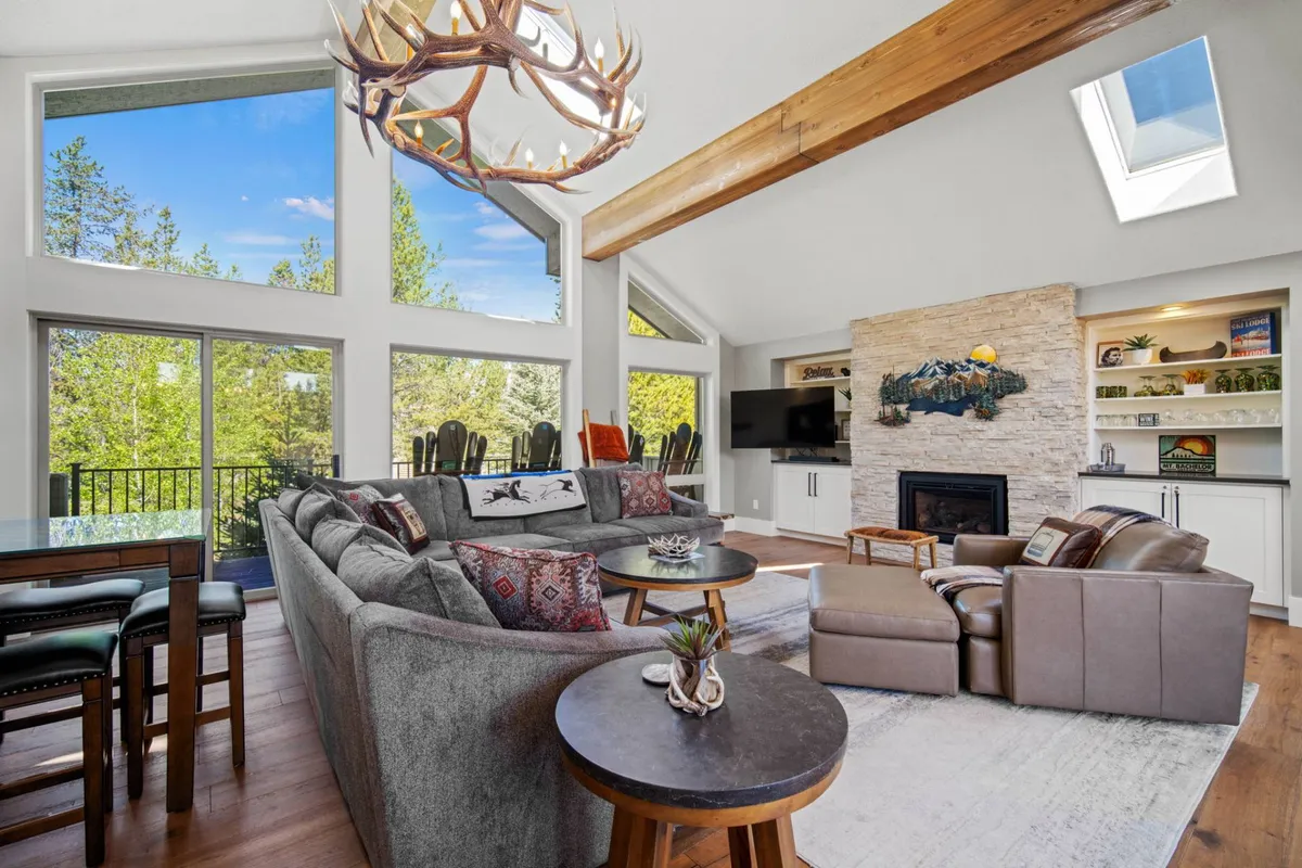 A welcoming living room featuring a large sectional sofa centered around a stone fireplace and illuminated by an antler chandelier. A wall of expansive windows reveals lush greenery outside, allowing sunlight to flood the space and highlighting the plush, contemporary furnishings.