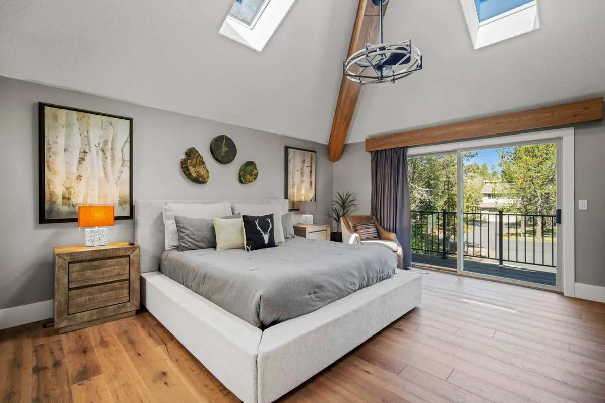 A serene bedroom with a large bed, skylights that bathe the space in natural light, a window with a view, striking artwork, and exposed wooden beams. The overall feel is cozy, modern, and luxurious.