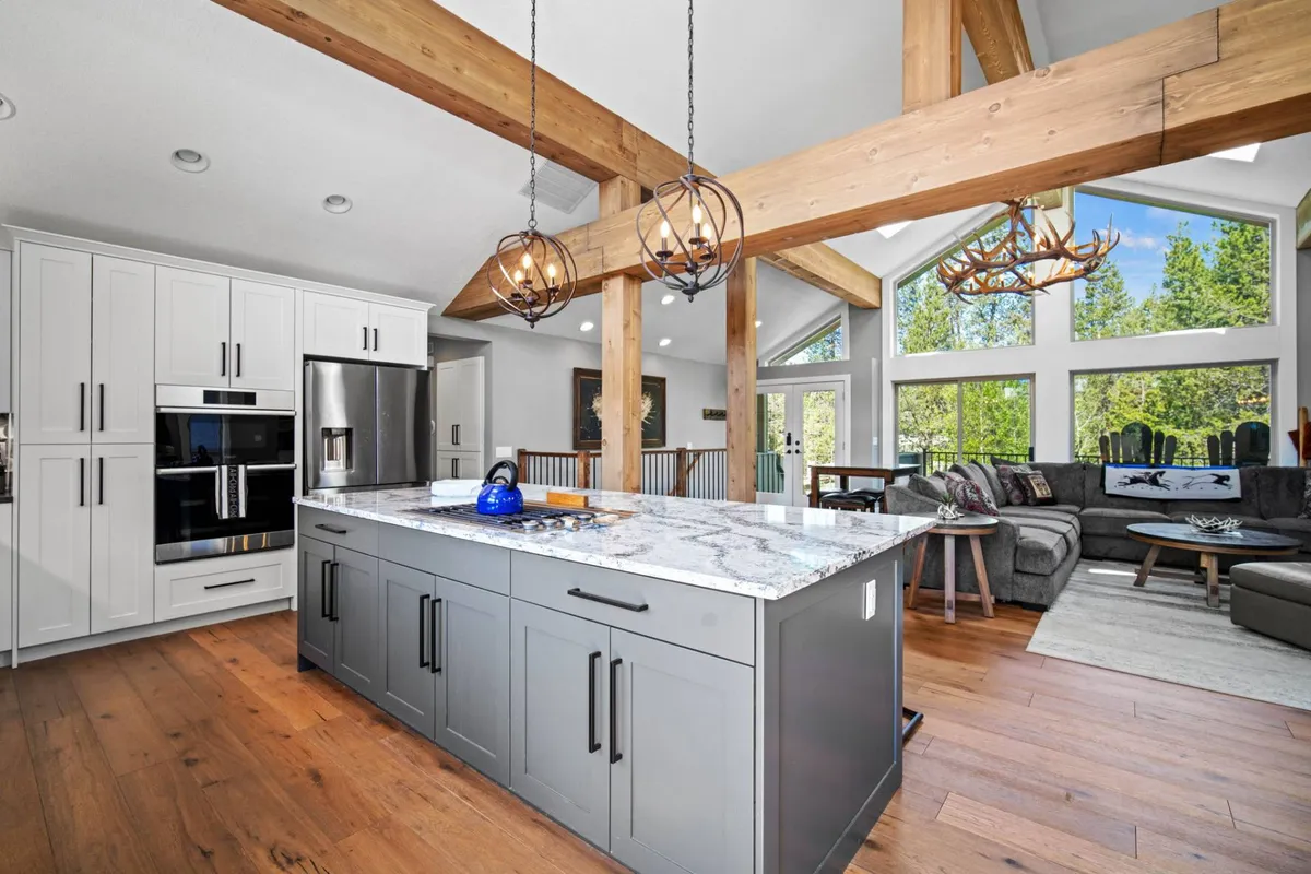 A bright and modern kitchen area featuring a central granite-topped island, warm wood beams, and large windows that allow natural light to pour in. The inviting setup is ideal for cooking, hosting, or enjoying morning rituals with family and friends.
