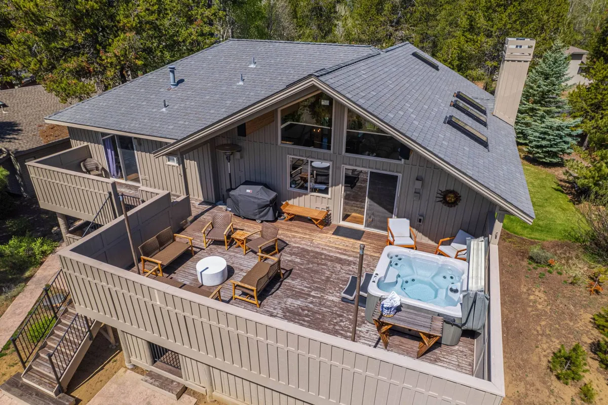 This image showcases an expansive wooden deck nestled among tall pines, prominently featuring a bubbling hot tub with steam rising and inviting outdoor lounge seating nearby. Large windows from the home’s interior reflect the golden hues of sunset, while the deck’s layout encourages relaxation and socializing. The setting conveys both tranquility and the luxury of soaking under the open sky, perfectly integrating natural surroundings with comfort.