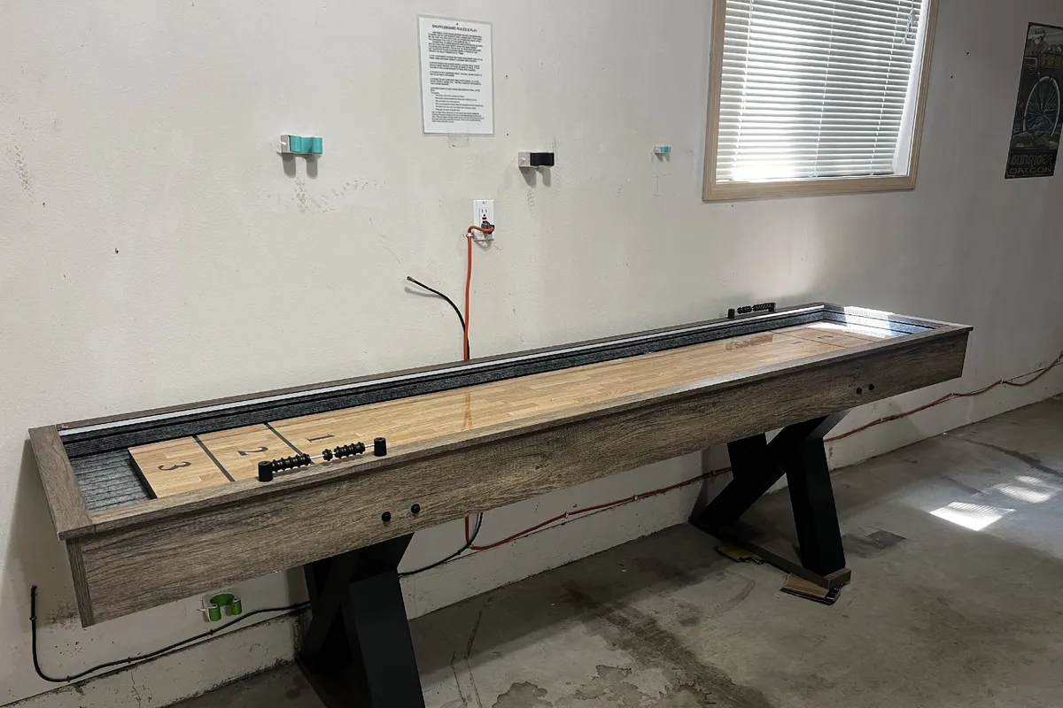 A shuffleboard table stretches along a minimally decorated wall with natural light streaming in from a nearby window. The clean lines and bright mood create an inviting atmosphere for casual competition and social play.
