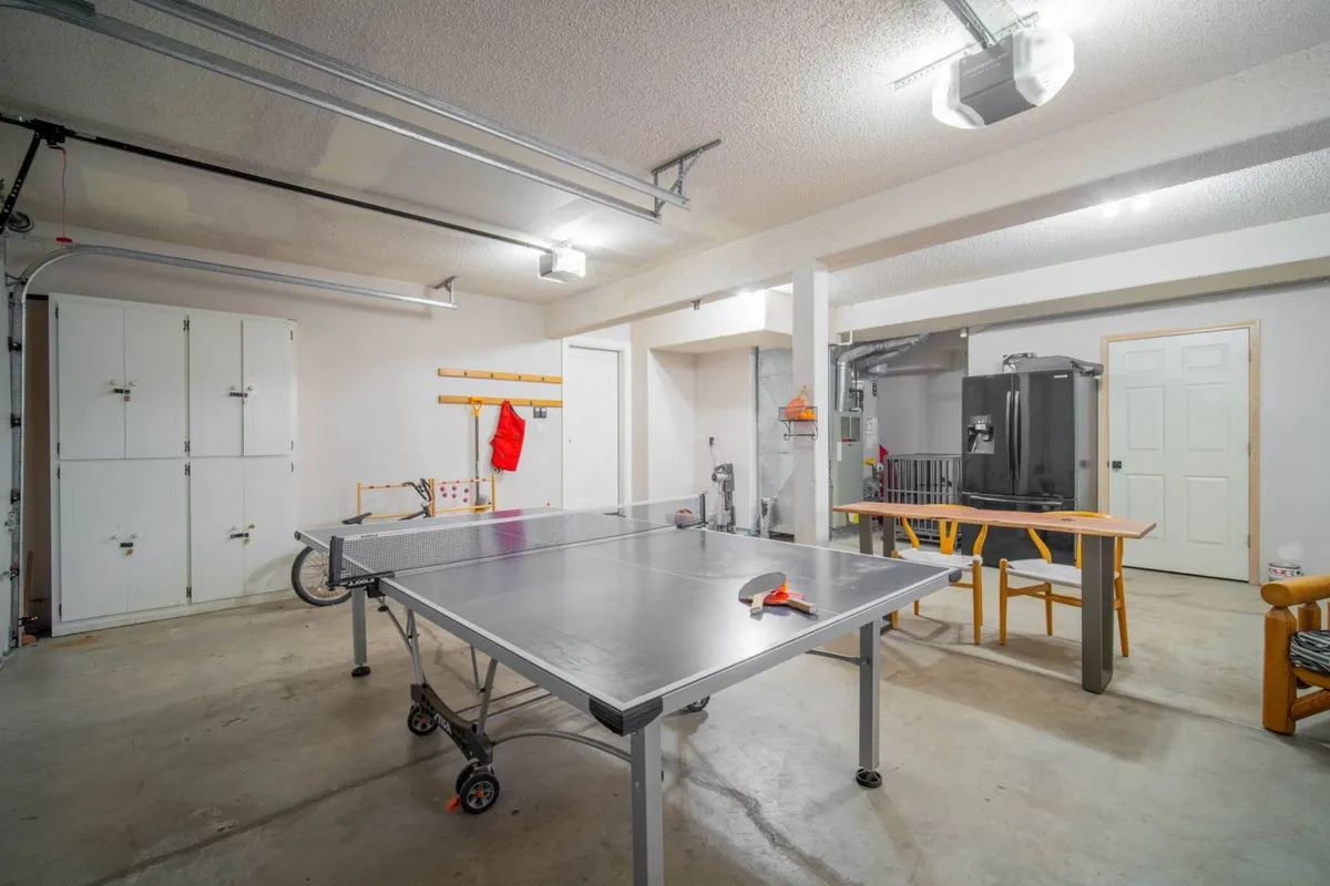 This image features a ping pong table positioned in a spacious garage setting, with storage cabinets and a work table visible along the walls. The casual layout suggests a multipurpose space ideal for games and gatherings. The doorway hints at easy access to other parts of the house, reinforcing the convenient location just off the main living area.