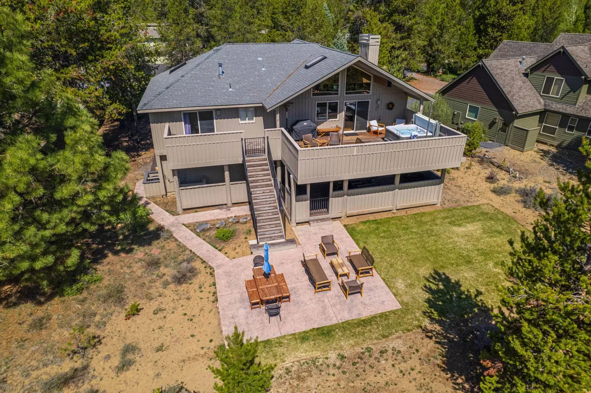 A broad deck stretches out with ample outdoor seating areas, surrounded by lush greenery. This space is clearly designed for outdoor dining and lounging, with comfortable chairs and a table inviting guests to gather, sunbathe, or enjoy meals together. The deck is bathed in soft natural light, reinforcing the idea of morning coffee or sunset views amidst nature.