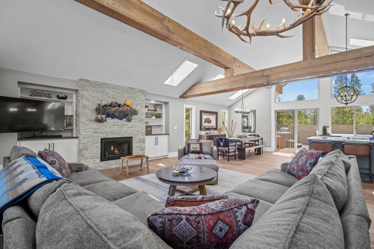 The image showcases a spacious and stylish open-concept living area, featuring a cozy fireplace, a plush large sectional sofa perfect for gatherings, an eye-catching antler chandelier, and an inviting dining area. Through expansive windows, tranquil views of the surrounding nature are visible, enhancing the sense of retreat and relaxation.